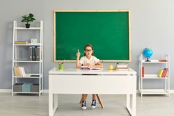 School child in glasses pointing finger up sitting at desk in front of green chalkboard. Cute student girl keen on learning playing in classroom, pretending to be a teacher and thinking of great ideas