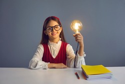 Portrait of a smart girl sitting at a school desk and holding a bright light bulb as a symbol of an idea. Concept of smart children, ideas, inspiration and new knowledge. Gray background.