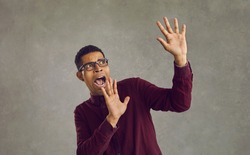 Frightened african american man screams in horror waving his arms on a gray background. Man with a look of fear on his face makes a frightened gesture with his palms as if trying to defend himself.
