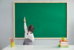 Back to school. Little schoolgirl writes on the school blackboard with chalk in the classroom.