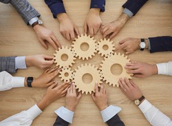 Team of business people join gearwheels. High angle, overhead view of circle of hands holding cogs on office table. Metaphor for good effective business system, cooperation, teamwork and efficiency