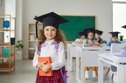 Portrait of a beautiful little curly elementary school graduate girl wearing an academic hat. Child with a book in his hands stands in the classroom and looks at the camera. Education concept.