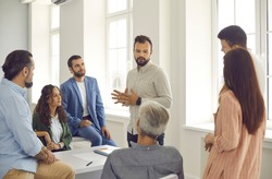Small group of diverse women and men listen to a confident male corporate business coach. Man advises clients, trains employees or explains the strategy of a marketing seminar. Staff training concept.