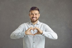 Handsome caucasian young man expresses a gesture of love while standing on a gray background. Smiling man in front of the camera shows his heart with his hands. Concept of romance and human emotions.