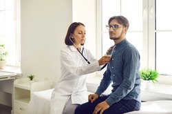 Young man visiting doctor for health exam. Serious middle aged female physician with stethoscope sitting on modern examination couch and listening to male patient's lungs or checking his heartbeat