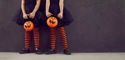 Halloween party banner. Cropped image of legs of two little girls with pumpkin candy bucket standing on black background. Children in black dresses and black and orange leggings stand near copy space