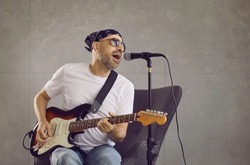 Portrait of male musician who sings song into microphone and plays electric guitar while sitting on gray background. Emotional middle-aged man dressed in rock style sings author's song. Studio shot.