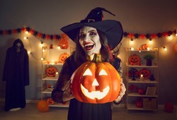 Horror night. Halloween portrait of spooky and evil witch with glowing jack-o-lantern in hands. Young woman in witch's hat with crazy and creepy smile looks into camera while standing in dark room.