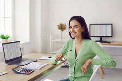 Portrait of beautiful happy female accountant sitting at her workplace in office. Smiling accountant or business analyst in casual clothes sitting in front of laptop , documents and calculator.