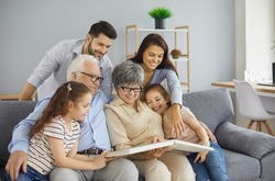 Happy big family grandparents with twin granddaughters and their parents browse the family photo album and share happy memories. Family gathered together in the living room. Family connection concept.