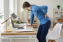 Tired man feels spasm and intense back pain as he stands up after working on computer in sedentary posture for long time. Unhappy stressed young employee has radiculitis or pinched nerve inflammation