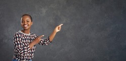 Smiling African American lady with short haircut promoting new product. Happy attractive young black woman in modern geometric pattern jumper points index fingers away at grey copy space background
