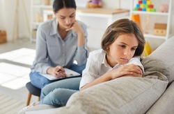 Frustrated little child at psychologist's office. Portrait of girl with sad face sitting on couch during therapy session. Therapist trying to help unhappy, resentful kid who has behaviour issues
