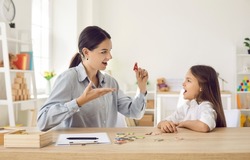 Female speech therapist working on dyslexic kid's problems and impediments. Happy child together with speech language pathologist sitting at a desk in modern office, playing fun game, learning letters