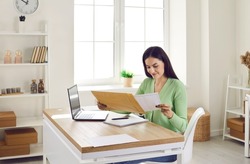 Business correspondence. Smiling woman receive postal mail with important document, invitation or bank message. Young woman working on laptop in her home office pulls out paper document from envelope.