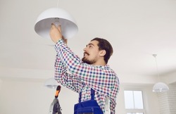 Electrician changing light bulb. Young man in overall work uniform standing on ladder in residential building, office, school or hospital and screwing LED lightbulb into modern white ceiling lamp