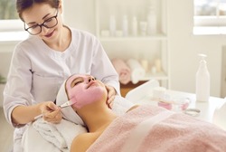 Happy woman getting facial treatment done by beautician in beauty salon or spa center. Beautiful young lady relaxing under soft towel and enjoying pink clay face mask for clear fresh rejuvenated skin