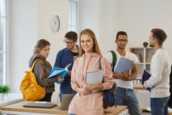 Portrait of smiling millennial teen girl student with backpack and book in university with mates. Happy young Caucasian teenager learner in school or college with groupmates. Education concept.