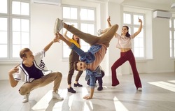 Flexible and energetic male dancer has fun with his dance group doing elements of breakdance movements. Young people in stylish youth casual clothes dance in bright dance hall with windows.