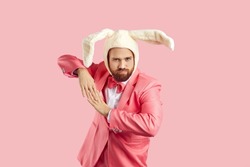 Rabbit costume party. Cheerful man dressed in funny fluffy hat with rabbit ears having fun isolated on pink background. Man in pink suit makes dance moves with funny expression on his face. Banner.
