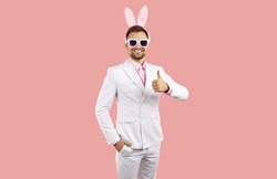 Handsome man with bunny ears wishing you Happy Easter. Happy attractive young guy in white stylish classic suit and funny plush rabbit ears standing isolated on solid pastel pink and showing thumbs up