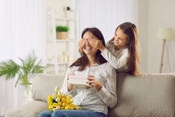 Happy young woman receives a present from her child on Mother's Day. Little daughter covers her mom's eyes as she gives her a gift and a bouquet of beautiful, fresh tulips. Surprise concept