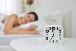 Alarm clock that is going to ring at seven in morning on background of woman sleeping in bed. Close up of alarm clock standing on table next to young woman sleeping sweetly in her bed. Selective focus