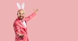 Funny man wearing bunny ears inviting you to Easter party. Happy young guy in pink suit and rabbit ears smiling and showing something on the right side on blank empty solid pink copy space background