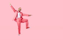 Cheerful young man in suit with suspenders makes funny movements on pastel pink background. Happy caucasian man in pink suit having fun and dancing near copy space. Full height. Banner.