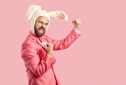 Happy Easter Bunny celebrating success. Funny cheerful confident ginger man wearing white fluffy rabbit ears and funky pink suit standing on pink background, fist pumping and shouting Yes, Yay, Hooray