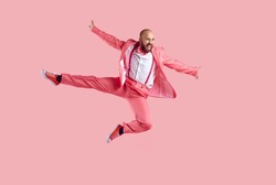 Happy confident young bald man jumping in the studio. Full body shot of a funny joyful male dancer wearing a party suit, bowtie and trainer shoes jumping high in the air isolated on a pink background