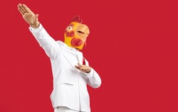Funny crazy man in white formal suit dancing chicken mask on his head isolated on red background. Man with rubber mask chicken or rooster having fun near copy space. Creative advertising banner.