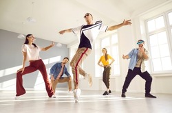 Expressive contemporary hip hop dance. Young male dancer performs hip hop movement on one leg during break dance lesson. Guy dances on surrounded by his dance friends in bright hall of dance studio.