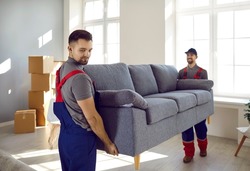 Two professional relocation service workers in overalls move sofa in customer's apartment. Movers carry sofa, cardboard boxes and assembling furniture. Moving and delivery company services.