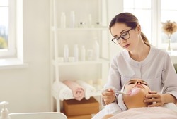 Beautician in glasses applying pink clay mask on woman's face. Calm beautiful young lady relaxing and enjoying facial procedures on spa day in modern beauty salon or spa center. Copy space background