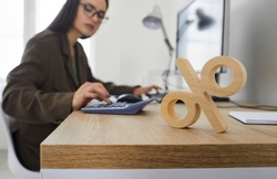 Corporate tax and interest rate. Close up of wooden percent sign standing on table on background of serious busy woman counting on calculator. Concept of interest calculation.