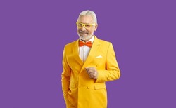 Studio shot of happy white haired bearded senior man wearing bright yellow suit, white shirt, orange bow tie and trendy glasses standing with his hand in pocket isolated on solid purple background