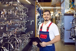 Portrait of a young man who sells faucets. Happy salesman in uniform standing in the aisle with modern water taps at a big hardware store or shopping mall