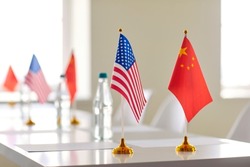 Flags of China and United States of America and papers for signing international agreement. Close up of small flagpoles with flags of two states on long white conference table without people.