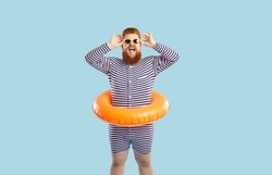 Funny funky cheerful man enjoying summer vacation isolated pastel turquoise background. Joyfully excited red-bearded guy in striped leotard and with inflatable swimming circle adjusts his sunglasses