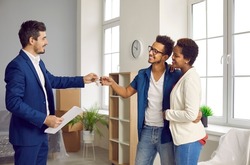 Smiling male real estate agent give keys to new house to excited multiethnic couple buyers. Realtor or broker congratulate multiracial spouse clients with home ownership. Realty and rental concept.
