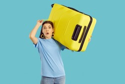 Surprised woman carrying her holiday luggage. Young lady standing isolated on blue background, holding yellow suitcase and looking at camera with funny shocked amazed facial expression. Travel concept