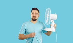 Man suffers from summer heat at home. Guy with broken air conditioner in his house using bad electric fan and sweating. Man in T shirt feeling hot and holding fan with sad face expression, studio shot