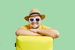 Happy man with suitcase who is waiting for summer trip isolated on light green background. Portrait of young smiling man in sunglasses and summer hat leans on yellow suitcase and looks at camera.