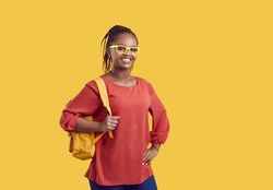Portrait of smiling millennial African American girl student in glasses with backpack isolated on yellow studio background. Happy young black female university or college graduate. Education concept.