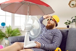 Funny sweaty chubby bearded man in swimsuit sitting at home, suffering from crazy summer heat, wiping sweat off forehead, holding electric fan, wishing for heatwave to stop and fresh breeze to blow