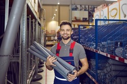 Happy man who works as a salesman at a hardware store where you can buy good quality tools and materials standing between shelves in one of the aisles, holding some gray PVC U drain pipes and smiling