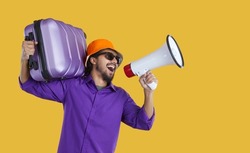 Excited man in purple shirt, orange hat and sunglasses standing on yellow studio background, holding travel bag and megaphone, advertising holiday sale, offering cheap tickets and fun summer vacation