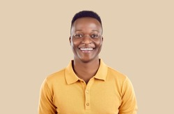 Studio portrait of happy African American man. Cheerful joyful handsome young guy in polo shirt standing on beige background, looking at camera and smiling
