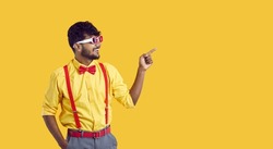 Happy young ethnic guy shows something on yellow copy space background. Cheerful Indian man in shirt, glasses, bowtie and pants with suspenders smiles and points his index finger to copyspace side
